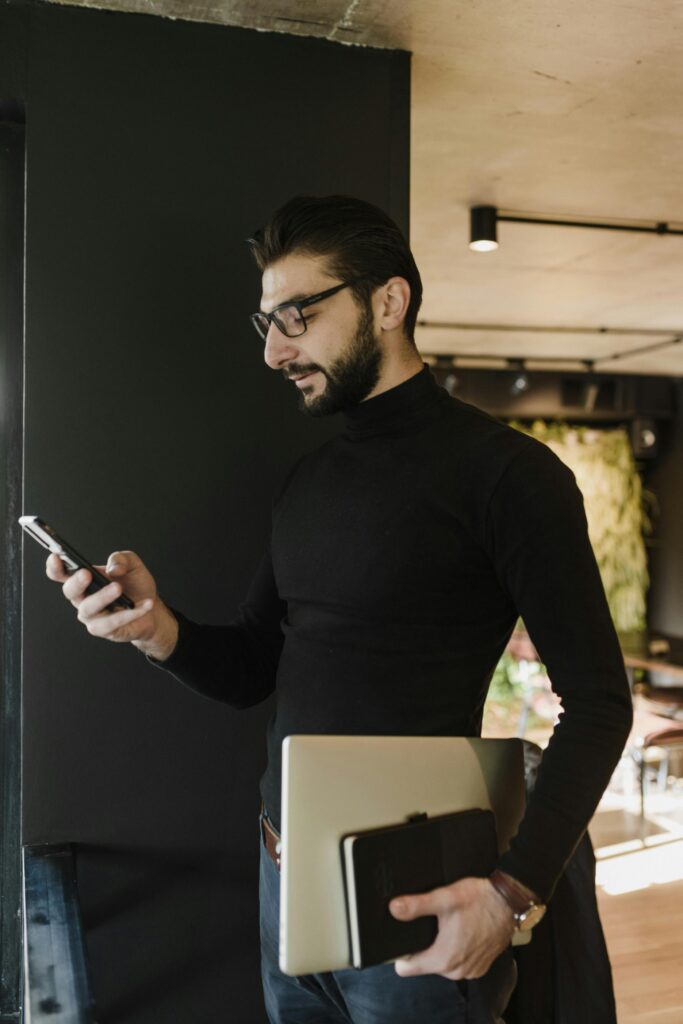 Young bearded man in black sweater using smartphone while holding laptop, standing indoors.