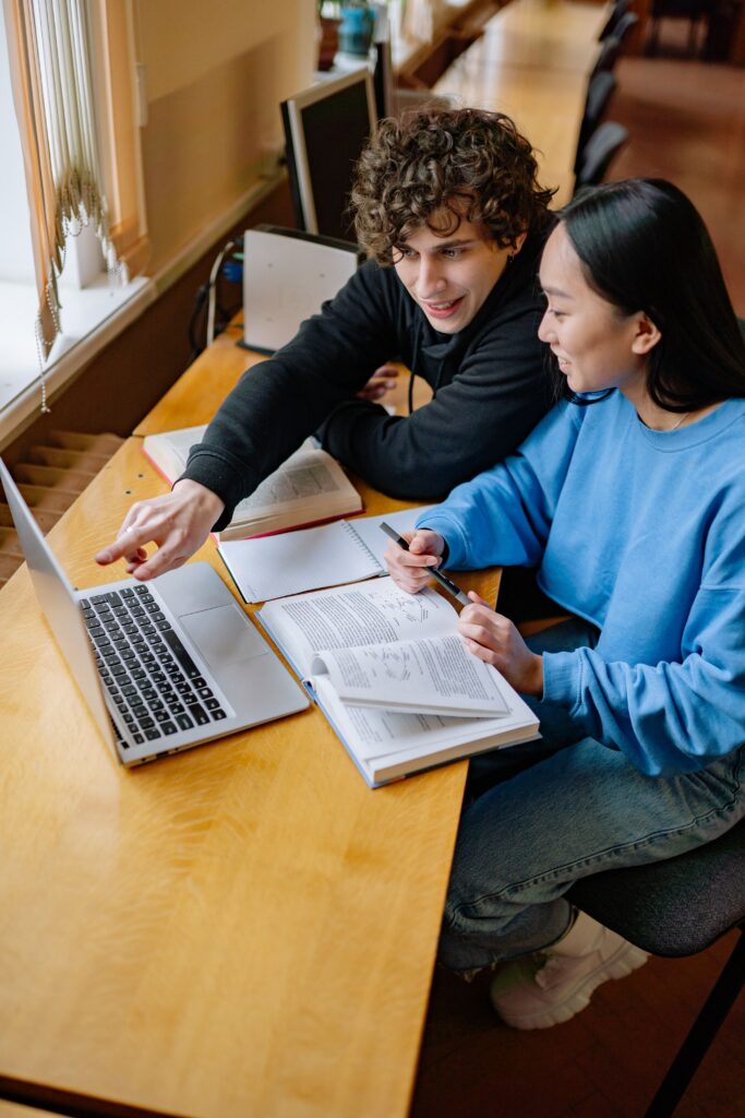 Two students working together on a laptop at a desk in a library, smiling and engaging.